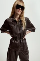 Chaqueta de efecto piel con cuello solapa, manga corta y cintura elástica. Presenta cierre frontal con cremallera y bolsillos decorativos en el frente.