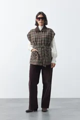 Chaleco de corte relajado con estampado a cuadros en tonos tierra, cuello camisero oscuro y cierre frontal con botones. Incluye un cinturón de la misma tela para ajustar a la cintura y bolsillos frontales.