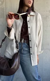 Chaqueta tipo sobrecamisa color beige, con cierre frontal de botones metálicos, cuello clásico y bolsillos frontales. Presenta un diseño de corte recto y mangas largas con puños abotonados.