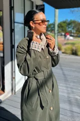 Trench reversible de corte clásico con cinturón ajustable. Un lado presenta un diseño liso en color verde militar, mientras que el reverso cuenta con un estampado a cuadros en tonos neutros. Posee cierre frontal con botones y cuello camisero.