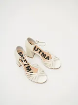 Sandalias de tacón cuadrado de 8 cm, confeccionadas en charol color crema. Presentan un diseño de tiras finas entrelazadas en la parte frontal y una correa ajustable con hebilla metálica en el tobillo.