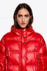 Chaqueta acolchada corta de tejido técnico brillante en color rojo, con cierre frontal de cremallera y botones de presión. Presenta capucha con borde de pelo sintético desmontable, bolsillos laterales con cremallera y puños de punto elástico en color rojo.