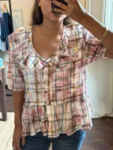 Camisa de manga corta con estampado a cuadros en tonos rosados, azules, amarillos y marrones. Presenta un cuello con volados y cierre frontal de botones. El bajo de la camisa tiene un detalle de volado.