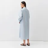 Vestido camisero midi celeste con finas rayas, cuello camisero, escote en V con botonadura frontal, bolsillos en el pecho, mangas largas anchas y aberturas laterales.