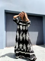 Vestido largo estilo túnica, color negro con estampado ornamental en blanco. Presenta cuello en V con botones y mangas largas amplias.
