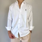 Camisa de algodón de corte regular, color blanco, con cuello clásico y cierre frontal de botones. Presenta un pequeño bordado oscuro en el pecho y mangas largas con puños doblados.