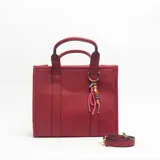 Cartera tipo tote de color rojo con textura granulada, doble asa superior y correa larga ajustable extraíble. Incluye un colgante decorativo con cuentas coloridas y tiras de cuero sintético en uno de los laterales.