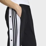Pantalon deportivo Adidas modelo Adibreak de corte holgado, color negro con franjas laterales blancas y las icónicas tres tiras en negro. Presenta botones a presión a lo largo de los laterales y logo de la marca en la parte inferior.