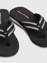 Ojotas negras con tiras de tejido trenzado en blanco y negro, y detalles distintivos de Tommy Hilfiger. La suela es de EVA con un tacón bajo.