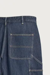 Pantalón jeans relaxed fit de mezclilla azul oscuro, con estilo carpintero que incluye bolsillos laterales y presillas de herramientas. Cuenta con costuras pespuntadas en contraste y cierre frontal con botón y cremallera.