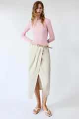 Falda midi de tiro medio color beige, con pliegues en la parte delantera, abertura frontal en el bajo y cinturón combinado a tono con hebilla metálica.