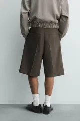 Bermuda de vestir color marrón oscuro, corte relajado y tiro alto, con pliegues frontales y cierre de botón y cremallera.