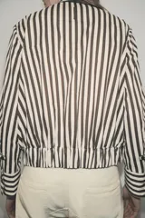 Camisa tipo bomber confeccionada en algodón, con diseño de rayas verticales en tonos marrón y blanco. Presenta cuello perkins, manga larga con puños, bolsillo de plastrón con solapa en el frente, bajo elástico y cierre frontal con botones.