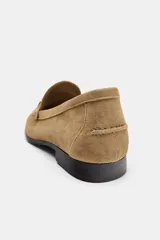 Mocasín de ante color marrón claro, con detalles de pespuntes en relieve, aplique metálico dorado en el empeine y suela plana con pequeño tacón.