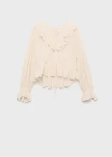 Blusa de gasa color crema con textura de pequeños puntos en relieve, cuello en pico con volantes, mangas largas abullonadas con puños acampanados y cierre frontal mediante lazos.