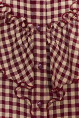 Blusa de manga larga con estampado de cuadros vichy en tonos burdeos y beige. Presenta cuello mao y grandes volantes fruncidos que cruzan el pecho y los hombros. Los puños son elásticos y fruncidos.