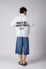 Camisa de manga corta a rayas beige y blancas, de corte boxy y liviana, con cuello camisero, cierre frontal de botones y pequeño texto bordado "BUILD IT BOLD" en el pecho.