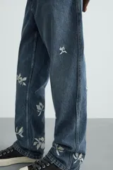 Jeans de corte relaxed fit en denim de algodón, color azul medio lavado. Presenta bordados florales en color blanco o crudo distribuidos en las piernas. Modelo de cinco bolsillos con cierre frontal de cremallera y botón.