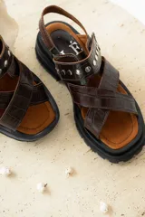 Sandalias de cuero marrón con textura de cocodrilo, diseño de tiras cruzadas con tachas metálicas plateadas y suela track negra de plataforma.