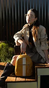 Mochila de cuero sintético color mostaza con diseño capitoneado. Presenta una franja vertical central en tonos blanco y beige, bolsillo frontal con cierre y correas regulables.