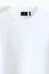 Remera blanca de manga corta, cuello redondo ribeteado y corte relajado con hombros caídos. Fabricada en punto liviano con fibra COOLMAX.