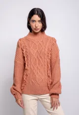 Sweater color crema de tejido trenzado con cuello alto, mangas largas abullonadas y detalle de flecos en el frente.