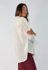 Camisa de algodón color blanco, con cuello redondo y detalle de volados en el cuello y puños. Presenta mangas largas con puños elásticos y un diseño de corte holgado con ruedo asimétrico, más largo en la parte trasera.