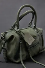 Cartera de cuero verde militar con acabado gastado, cuerpo blando, asas de mano, bolsillos frontales con cierre y tiras colgantes.