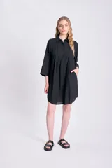 Vestido camisero corto negro, de manga tres cuartos, con cuello camisero y detalle de tablas verticales en el frente. Presenta bolsillos laterales y un calce holgado.