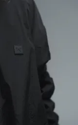 Campera rompevientos negra de calce oversize, confeccionada en microfibra técnica ligera con acabado semi-mate. Presenta capucha integrada, cierre frontal técnico y bolsillos tipo ojal. Destaca por su sistema de frunces elásticos regulables mediante tancas a lo largo de las mangas y el ruedo, permitiendo ajustar el volumen de la prenda.