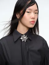 Camisa de corte clásico en color gris oscuro, con cuello camisero, mangas largas con puños abotonados y cierre frontal con botones. Incluye una corbata a tono con un broche decorativo en forma de flor metálica en el cuello.