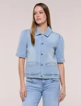 Camperita de denim color celeste con efecto lavado suave, cuello camisero clásico y cierre frontal de botones metálicos. Tiene mangas cortas con fruncido en los hombros, bolsillos delanteros bajos y costuras estructuradas.