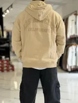 Canguro oversize beige con capucha y cordones ajustables, bolsillo tipo canguro y pequeño bordado en el pecho.