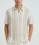 Camisa de hombre confeccionada en 100% algodón con textura, de manga corta y cuello inglés. Presenta un diseño de rayas verticales en tonos beige, blanco y marrón, con cierre frontal de botones.