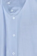 Camisa de algodón color celeste, con cuello alto y lazo integrado en el mismo tejido. Presenta cierre frontal con botones, manga larga con puños y un bolsillo frontal en el pecho. Diseño de corte recto.