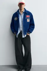Cazadora estilo varsity confeccionada en pana de algodón azul, con interior ligeramente acolchado. Presenta cuello solapa, cierre frontal con botones a presión, bolsillos laterales de vivo y acabados en rib a rayas en puños y cintura. Incluye múltiples parches bordados decorativos en el frente.