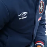 Campera tipo bomber azul marino con cierre frontal de botones a presión. Presenta el logo de Umbro bordado en el pecho derecho, el escudo del Club Nacional de Football en el pecho izquierdo, el año 1899 bordado debajo del escudo y detalles con los colores del club en cuello, puños y ruedo. Incluye parches laterales conmemorativos.