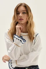 Blusa blanca de algodón con cuerpo de jersey y mangas de popelina. Presenta detalles de punto smock en puños y cuello, adornados con delicados bordados en azul y dorado.