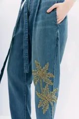 Pantalón de denim azul de tiro alto con cintura estilo paper bag y lazo a tono. Presenta un bordado elaborado con lentejuelas y piedras en forma de palmeras en la parte lateral de la pierna.
