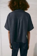 Camisa crop efecto denim color negro, con cuello solapa, escote en V, mangas cortas con trabilla y bolsillos frontales de parche. Cierre frontal con botones.