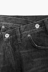 Jeans de tiro alto, color gris oscuro lavado, con pierna ancha extra larga (full length) y diseño de cintura cruzada asimétrica con cierre frontal de cremallera y botones metálicos.