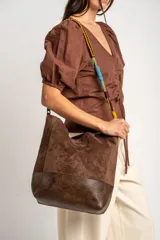 Bolso tipo shopper de tamaño maxi, confeccionado en material efecto gamuza marrón con base en cuero sintético oscuro. Cuenta con una correa ajustable y desmontable trenzada en varios colores.