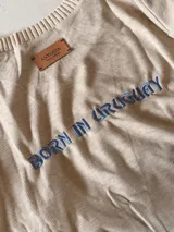 Sweater de punto color beige con cuello redondo y diseño de escudo bordado en el frente. Presenta un corte holgado y mangas largas.