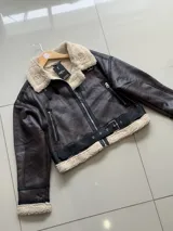 Campera tipo gamulán de cuero sintético en color marrón oscuro, con interior y cuello de corderito sintético en tono crema. Presenta cierre frontal, correas ajustables en el cuello y puños con terminación en corderito.