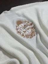 Ruana de tejido de punto con textura tipo panal en color blanco, con un escudo nacional bordado en hilo color dorado en el centro.