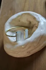 Pulsera rígida dorada con dos piedras rectangulares de color blanco nacarado en los extremos.