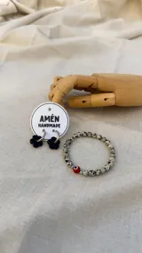Combo de accesorios compuesto por un par de aros de acero quirúrgico con dijes de flor de vidrio negra y una pulsera elástica de piedras naturales jaspe dálmata con un dije central de ojo turco.
