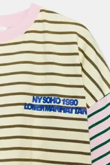 Camiseta oversize con diseño bicolor a rayas horizontales, combinando un lado en tonos rosado y verde oscuro, y el otro en beige y marrón. Cuenta con cuello redondo, mangas cortas amplias y texto bordado en azul en el pecho.