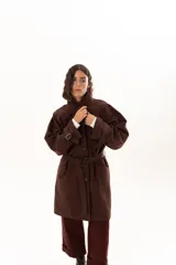 Trench color chocolate con cuello alto, cierre frontal con botones y cinturón ajustable en la cintura. Presenta mangas largas con puños ajustables mediante tiras y hebillas, y bolsillos frontales.