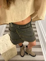 Minifalda cruzada tipo portafolio de denim color verde oscuro/gris, con bordes deshilachados en capas superpuestas y ajuste lateral con hebilla y ojalillos metálicos.